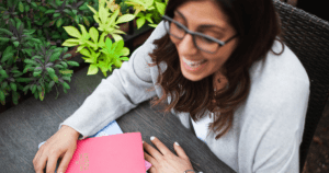 Happy, bespectacled woman at a table or desk that is beside a green plant. Her hands are resting on folders that are on the desk. Perhaps listening to someone